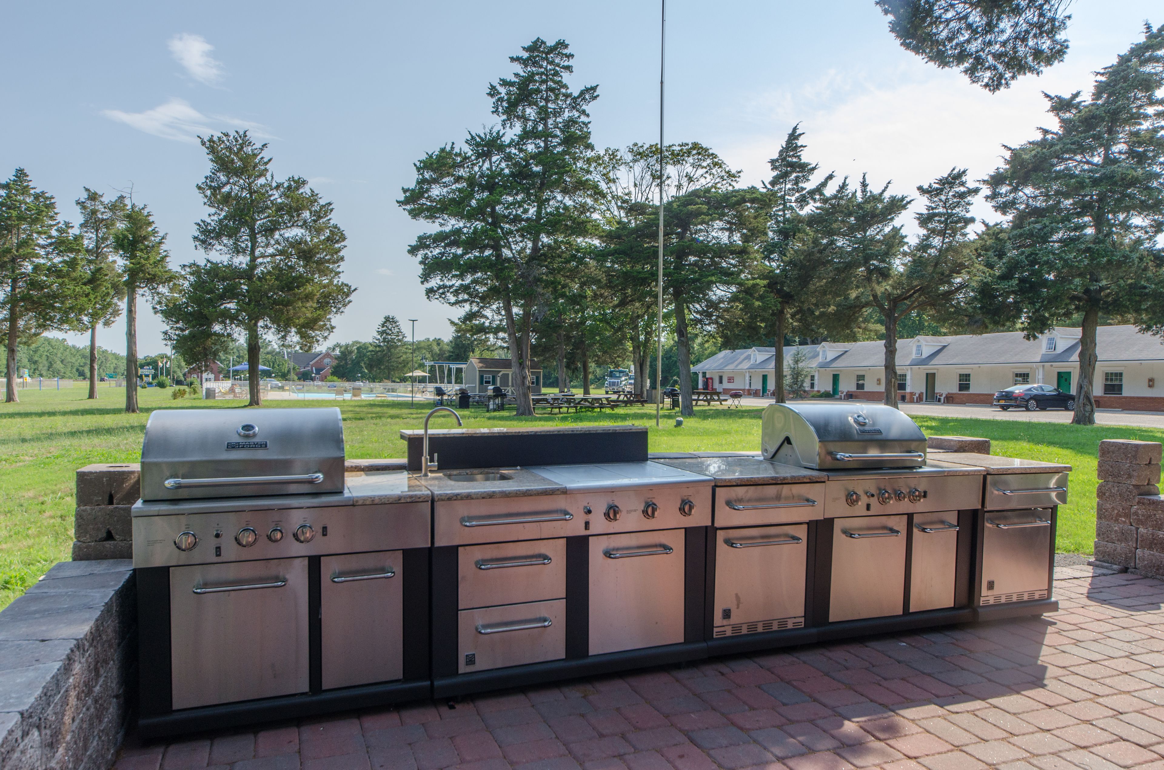 A large, stainless steel outdoor kitchen with multiple grills and sinks, set against a backdrop of trees and a grassy area.