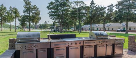 A large, stainless steel outdoor kitchen with multiple grills and sinks, set against a backdrop of trees and a grassy area.