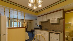Cozinha compacta com micro-ondas, fogão e pia. Há uma área de refeições com mesa e cadeiras. O quarto tem paredes amarelas e uma janela com cortinas.