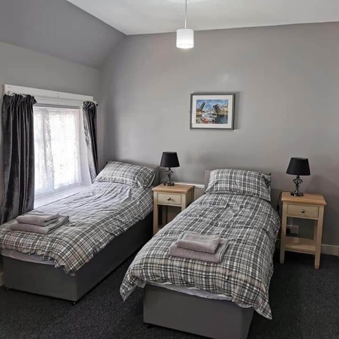 A room with two beds, each with a matching nightstand and lamp. There is a window with curtains, a framed picture on the wall, and a ceiling light.