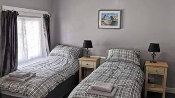 A room with two beds, each with a matching nightstand and lamp. There is a window with curtains, a framed picture on the wall, and a ceiling light.