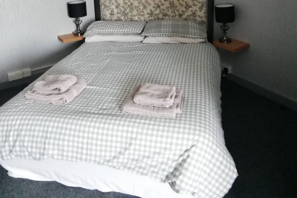 A neatly made bed with checkered bedding and two pillows, flanked by bedside tables with lamps, in a room with grey walls and framed plant artwork.