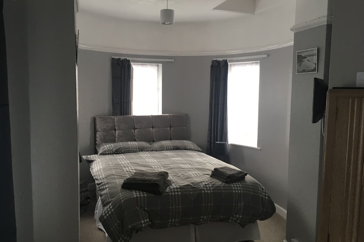 A bedroom with a bed, two windows, a ceiling fan, and a wooden wardrobe.