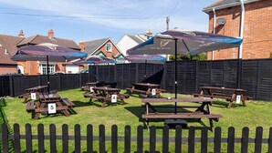 An outdoor seating area with numbered picnic tables and umbrellas, surrounded by a wooden fence and brick buildings in the background. - The Old Castle (Weymouth)