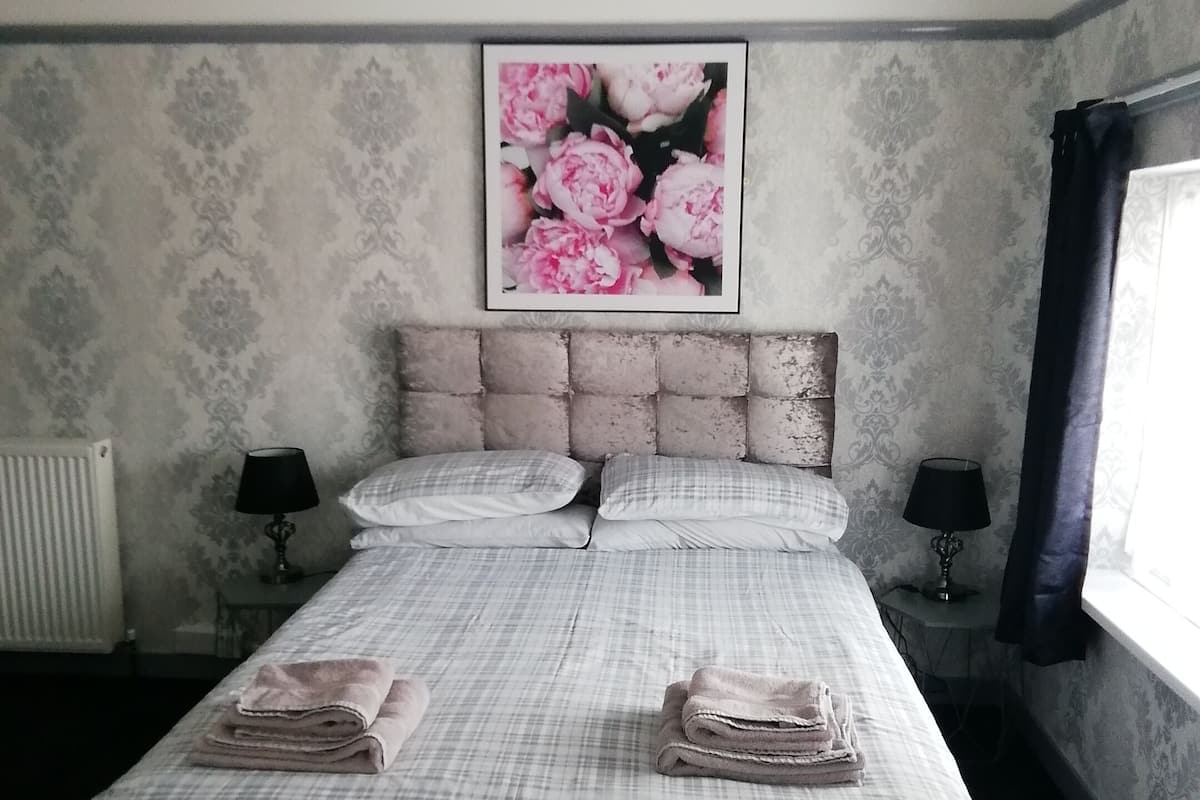 A bedroom with a bed, two bedside tables with lamps, a radiator, a window with curtains, and a floral wall hanging.