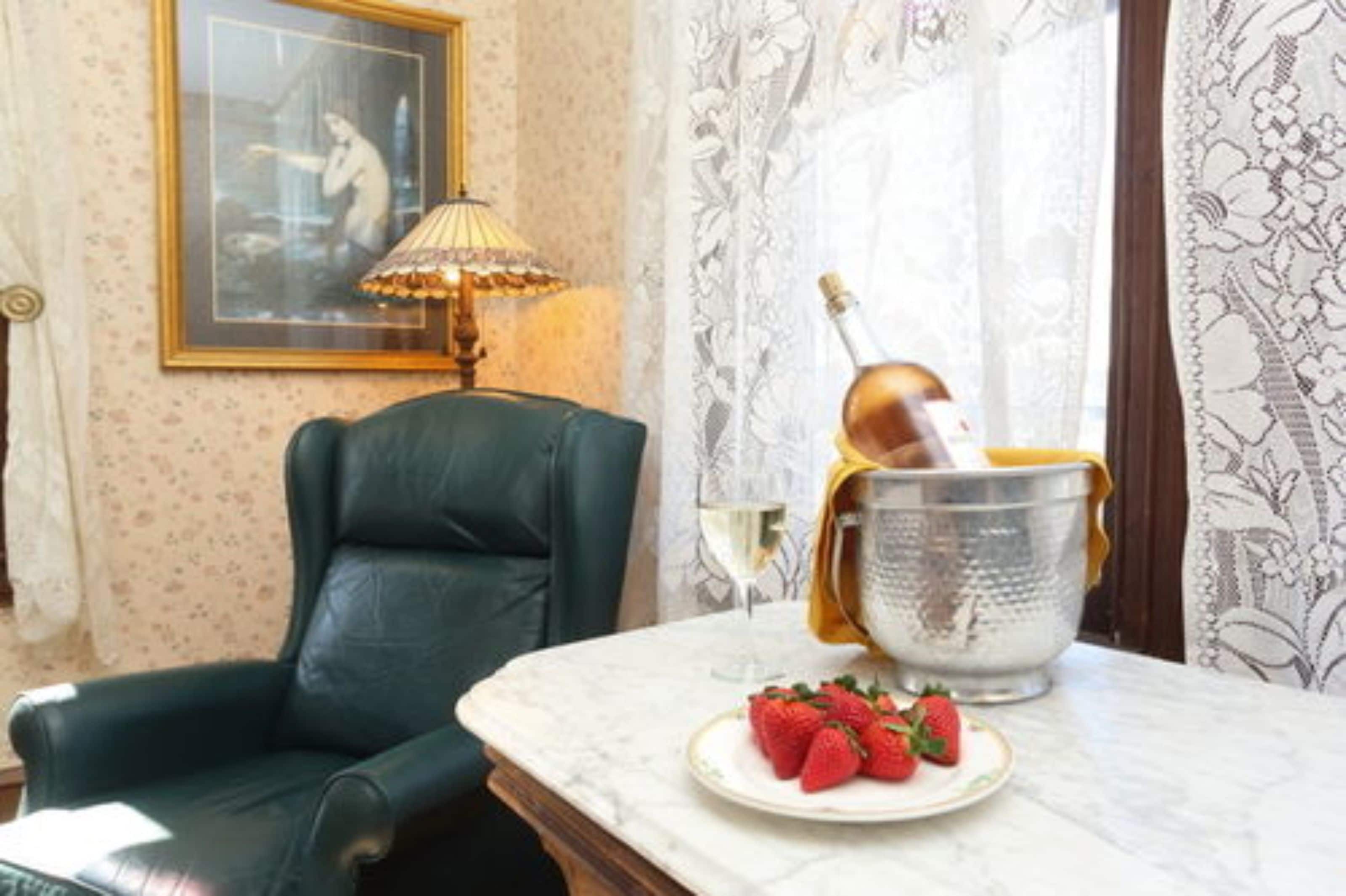 A room with a green leather chair, a table with a plate of strawberries, a bottle of champagne, and a lamp.