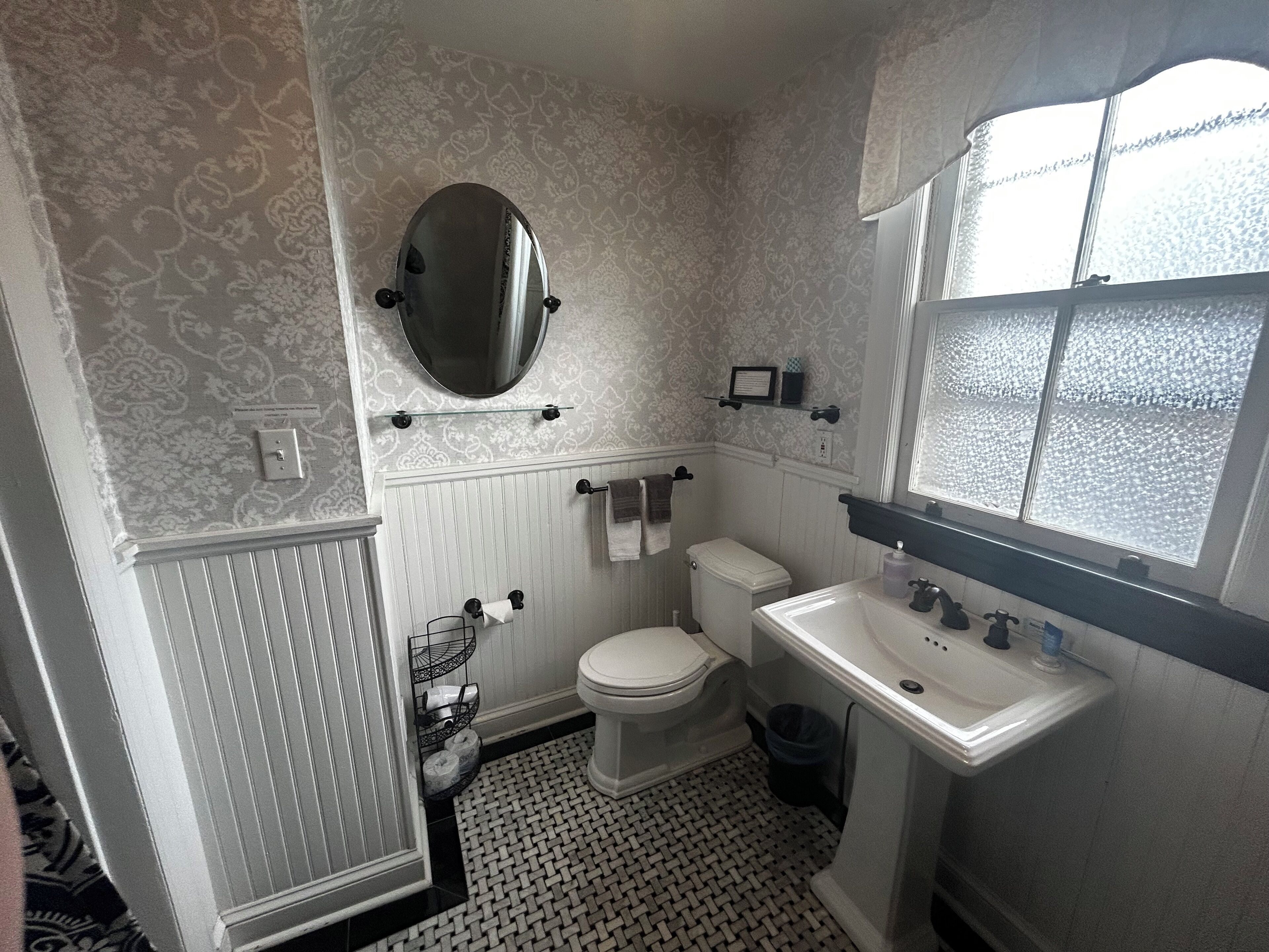 A bathroom with a toilet, sink, and mirror. There is a patterned wallpaper and a window with frosted glass.