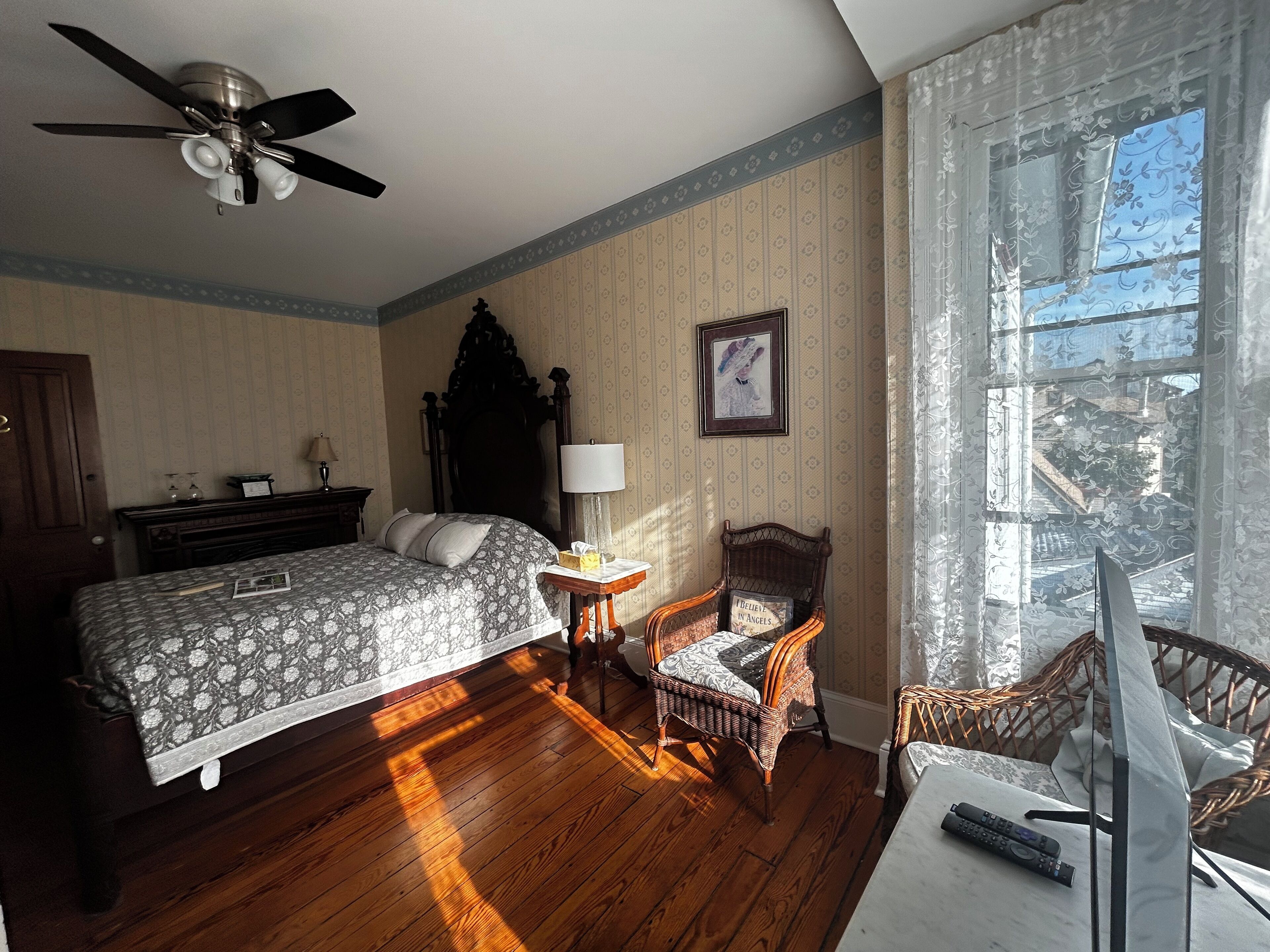 Une chambre à coucher comprenant un lit, une chaise, une table de chevet, une lampe, un ventilateur de plafond et une fenêtre avec des rideaux.