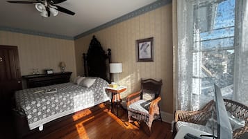 Une chambre à coucher comprenant un lit, une chaise, une table de chevet, une lampe, un ventilateur de plafond et une fenêtre avec des rideaux.