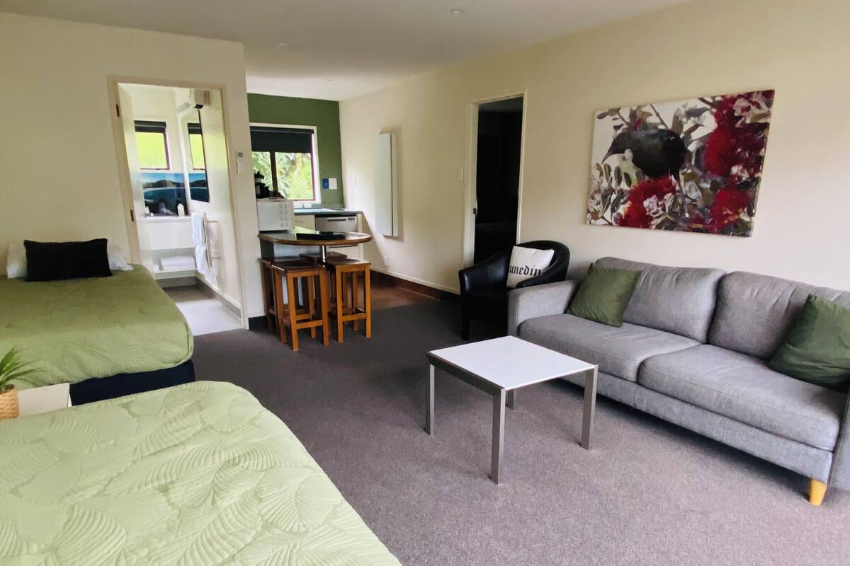A room with two beds, a sofa, a small table, and a chair. There is a kitchenette in the background.