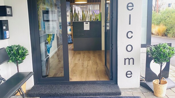 A reception area with a 'Welcome' sign, a reception desk, a bench, and a welcome mat outside. - Commodore Motels (Dunedin)
