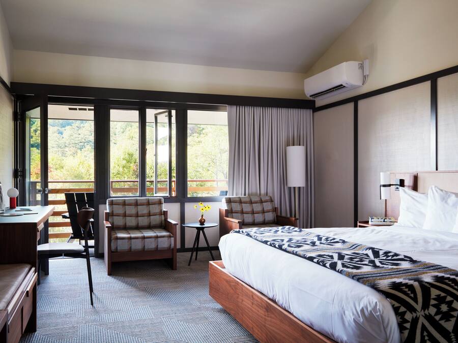 A hotel room with a large bed, a desk with a chair, a small table with a lamp, and a view of the outdoors through large windows.