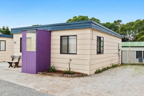 A small, single-story building with a beige exterior and a purple door. It has two windows and a small garden area with plants.