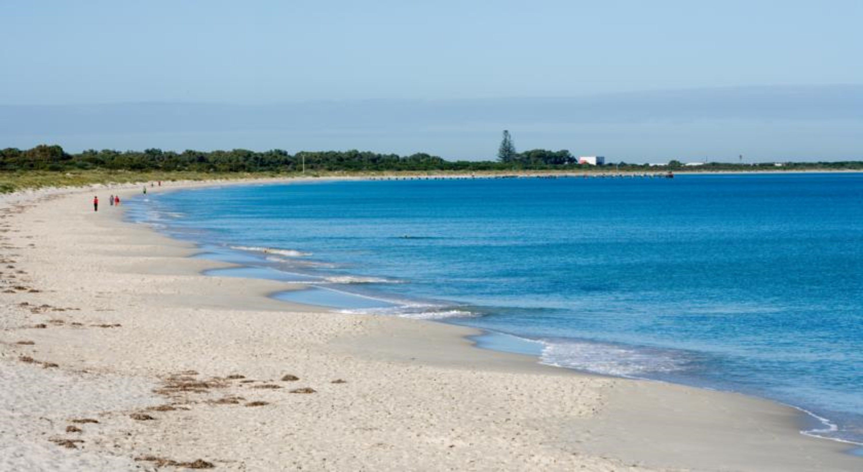 A beach with clear blue water, sandy shore, and a distant shoreline with greenery and buildings.