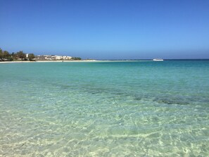 A clear view of a beach with shallow turquoise water, a distant boat, and a coastline with buildings and trees.