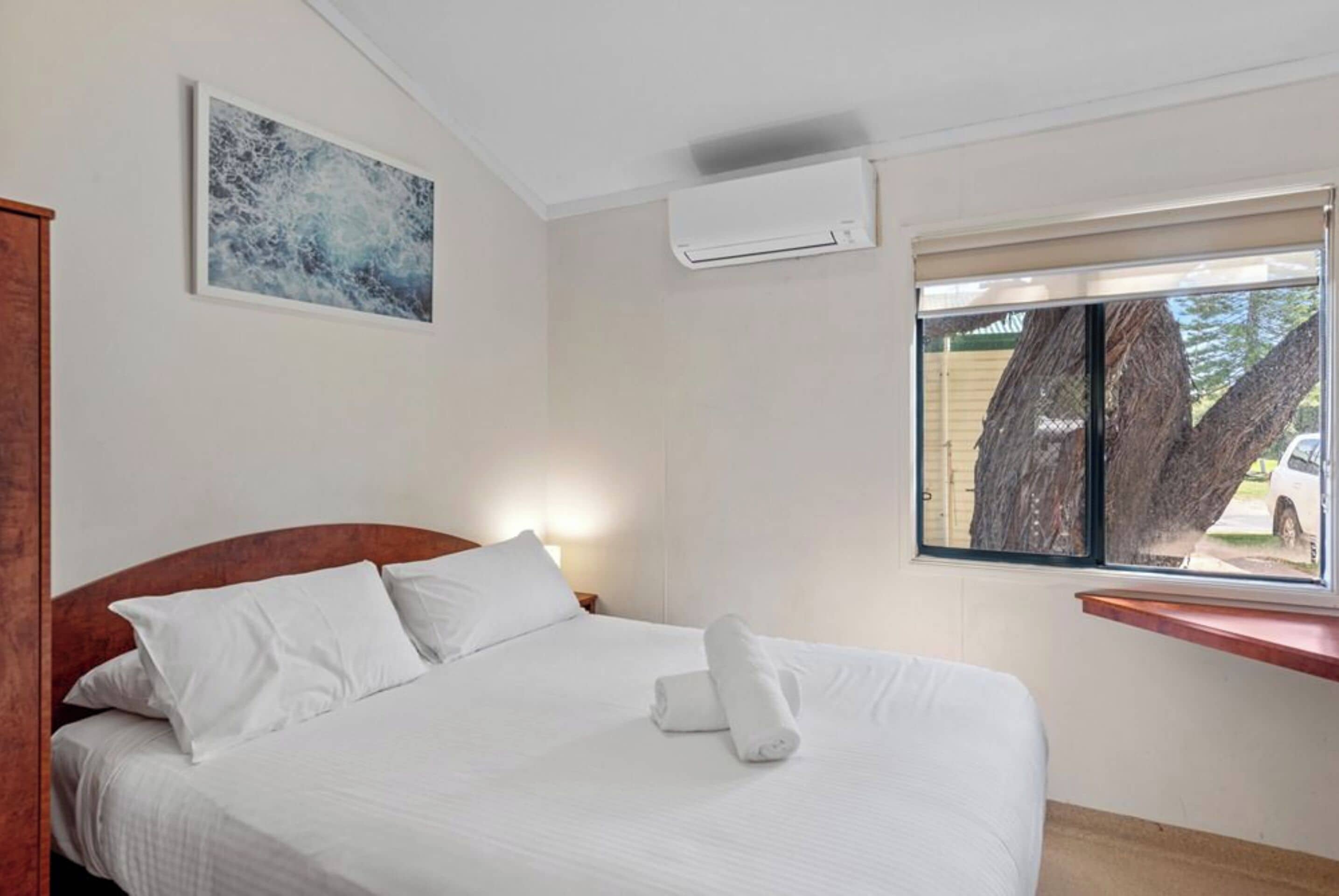 A neatly made bed with white linens and pillows, a wall-mounted air conditioner, a window with a view of trees and a parked car, and a framed picture on the wall.