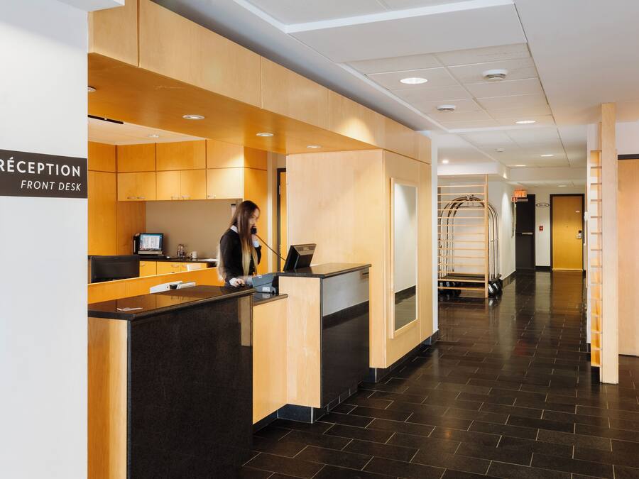 Una reception con un cartello che riporta 'RECEPTION FRONT DESK', una donna in piedi dietro e una scala che conduce a un piano superiore.