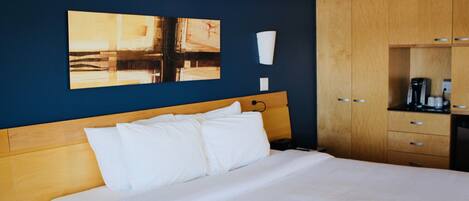 Habitación de hotel con una cama, cabecera de madera, un cuadro en la pared, una taza blanca y una cafetera.