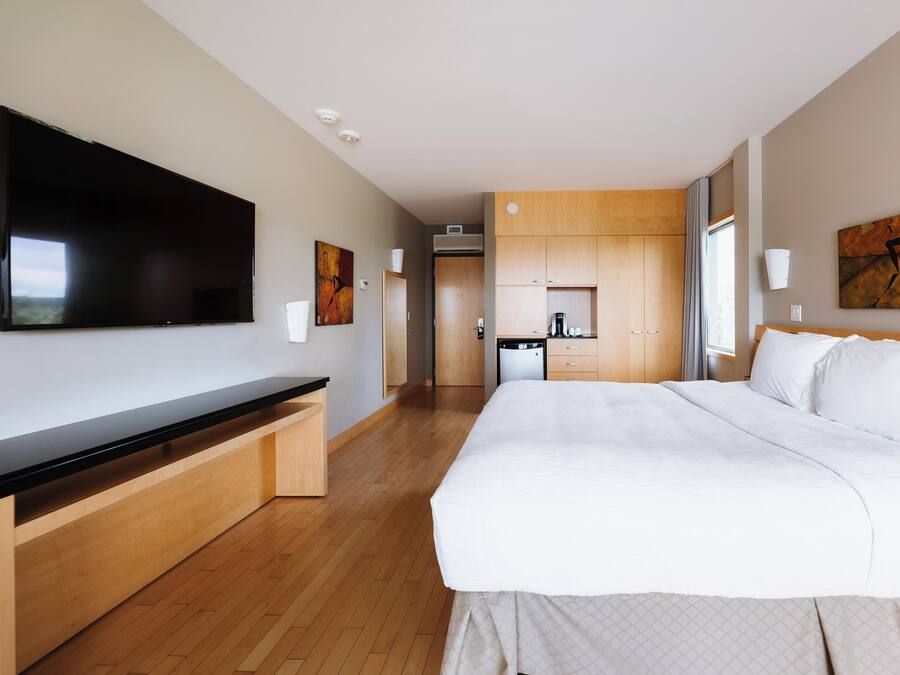 A modern hotel room with a large bed, a flat-screen TV, a wooden bench, a kitchenette, and a painting on the wall.