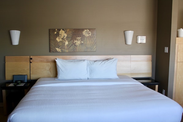 Habitación de hotel con una cama grande, cabecera de madera, dos mesitas de noche con lámparas, un escritorio pequeño con una laptop y una obra de arte colgada en la pared.