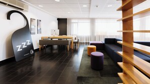 A modern hotel lobby with a large black wall art piece, a wooden reception desk, a seating area with a sofa and ottoman, and a large window with blinds. - Hôtel Le Dauphin Saint-Hyacinthe (Saint Hyacinthe)