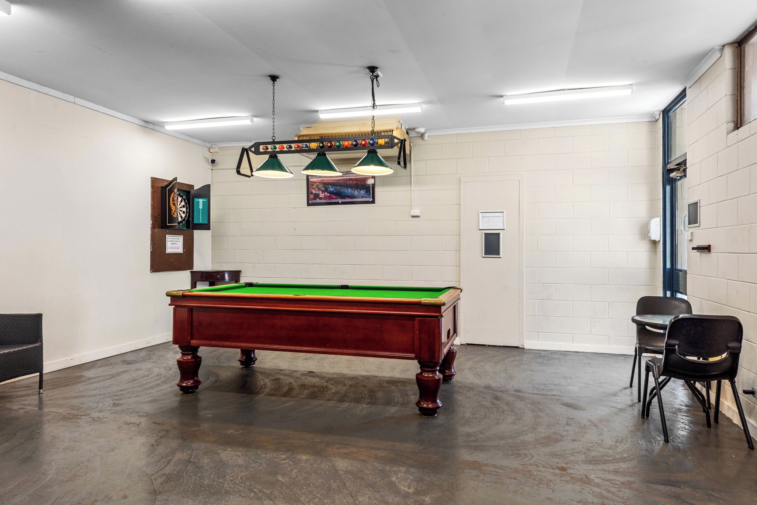 Una mesa de billar en una habitación con pared de ladrillo, una puerta y dos sillas.