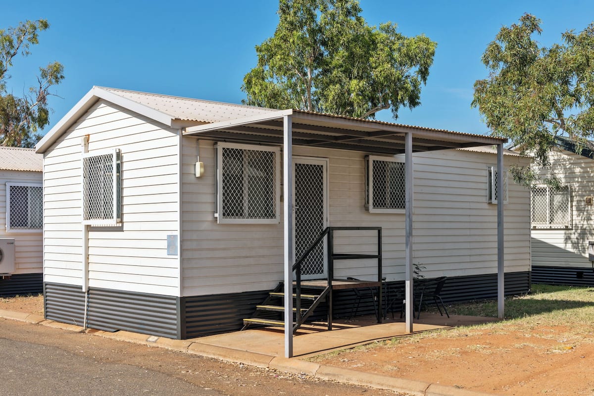 A small, single-story mobile home with a covered porch, a set of stairs, and a fenced area.