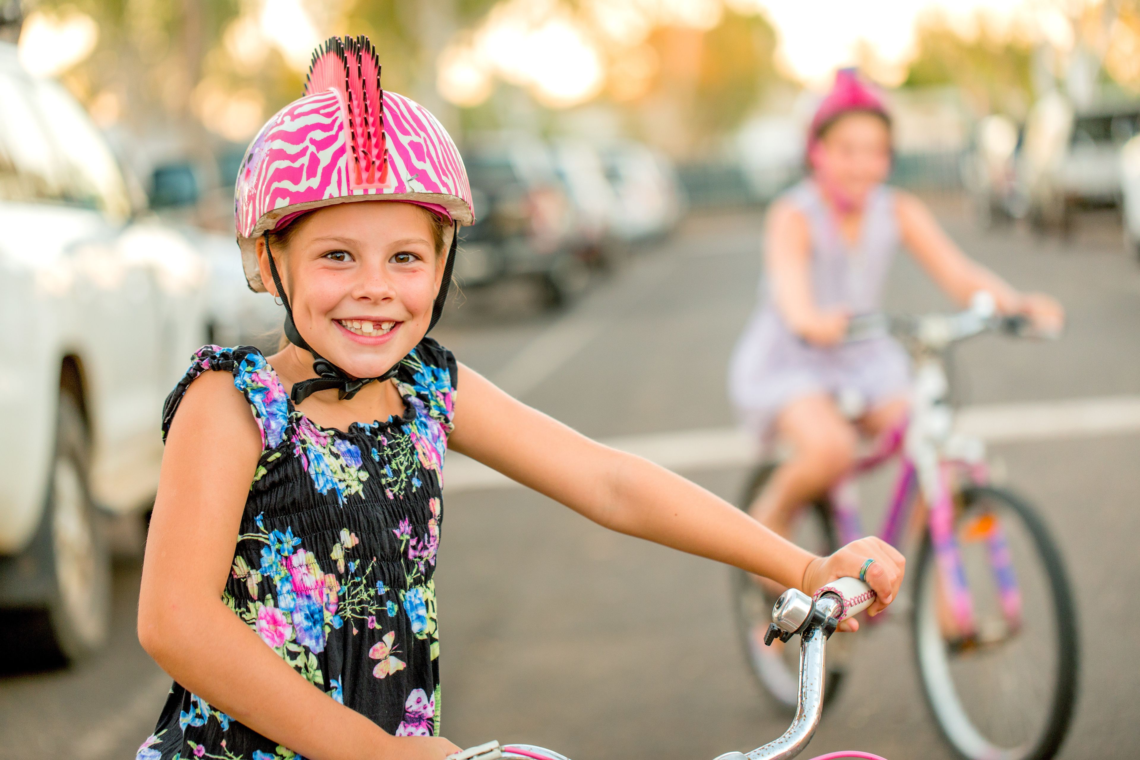 Dos niños en bicicleta, uno con casco y la otra con un vestido, en una calle con coches aparcados.