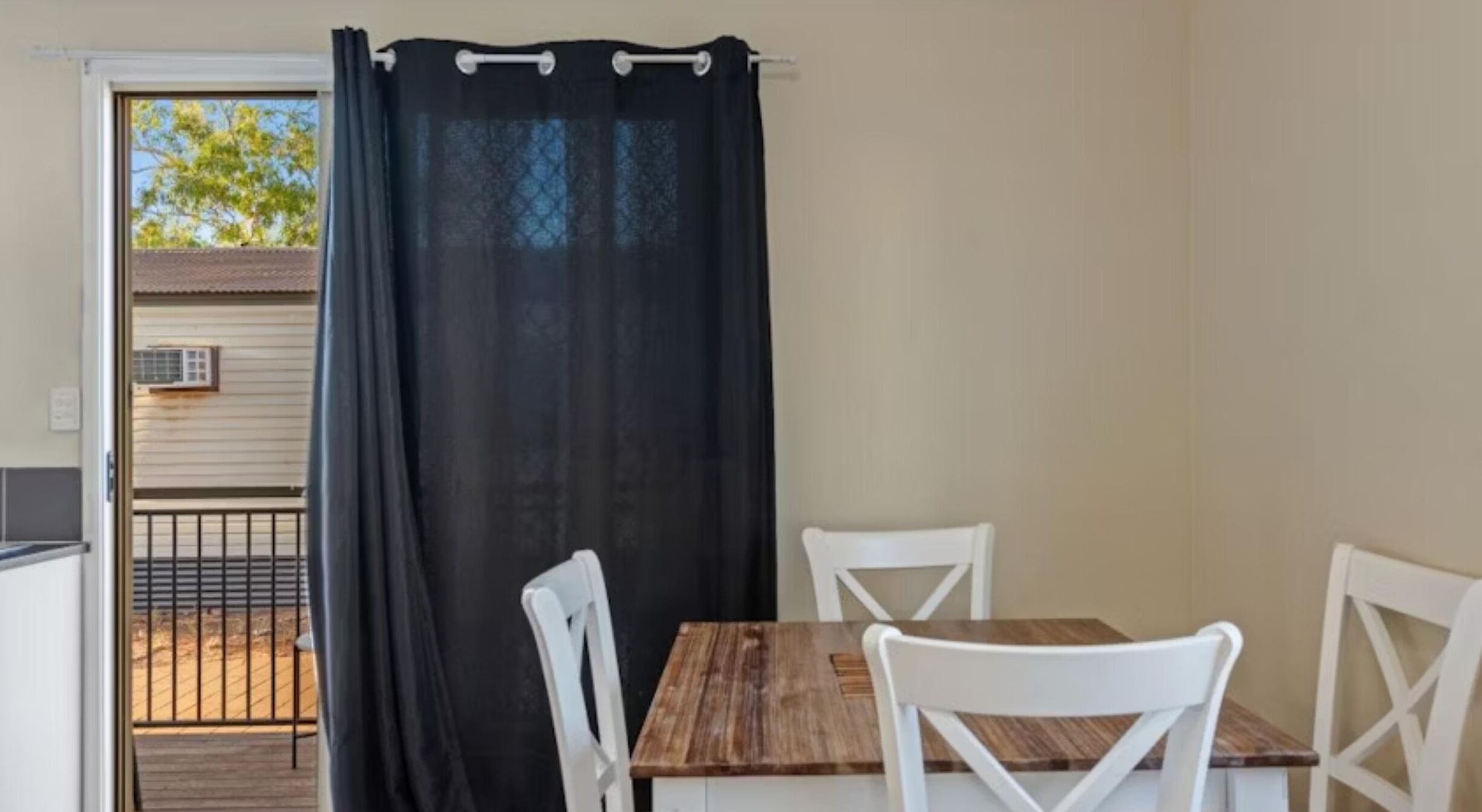 A small dining area with a wooden table and white chairs, a black curtain, and a view of a house through a glass door.