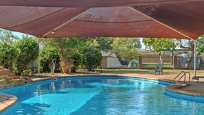 Una piscina con zona sombreada, rodeada de árboles y una valla.