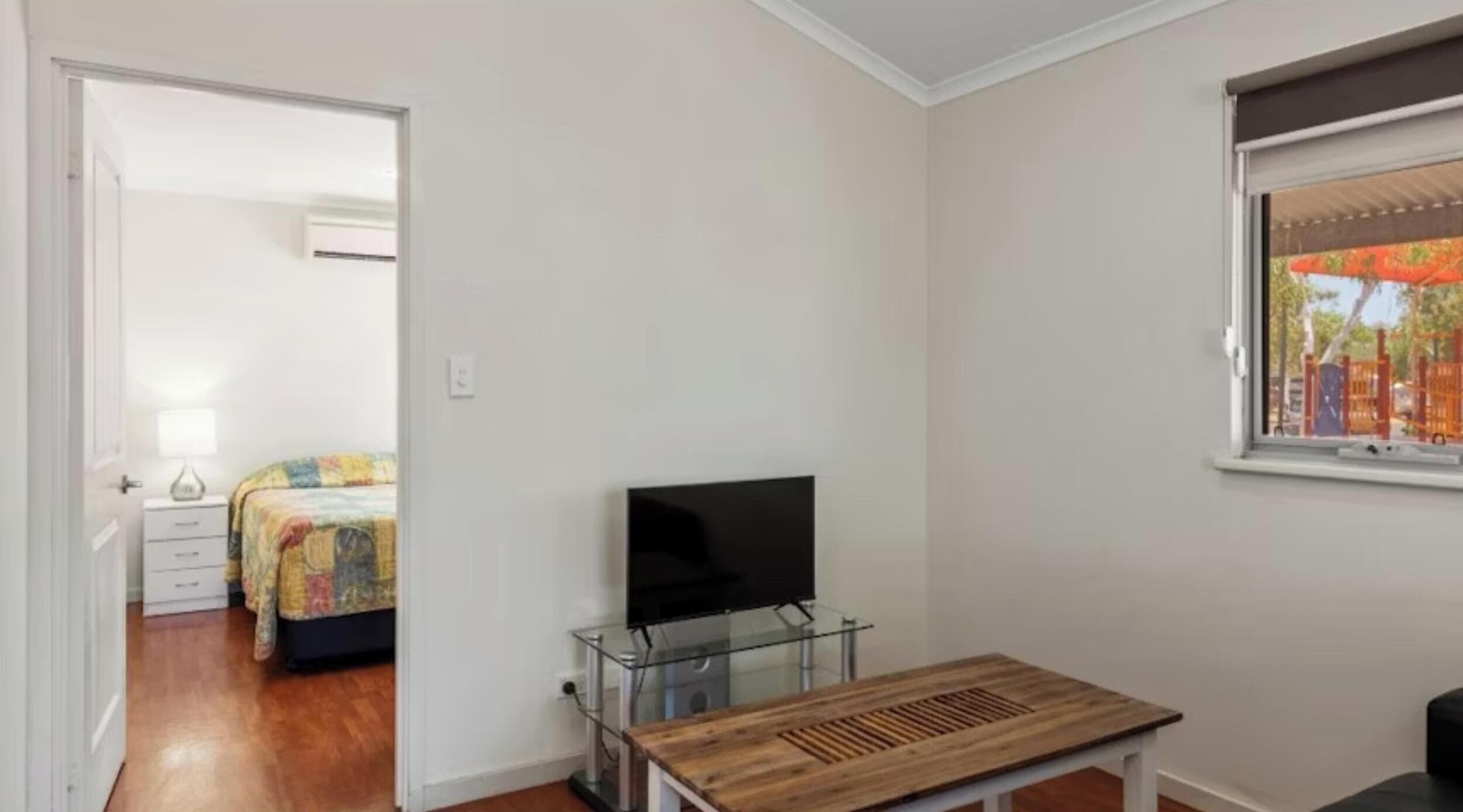 Un dormitorio con cama, televisor sobre un pedestal de vidrio, mesa de madera y ventana con vista a árboles y un parque infantil.