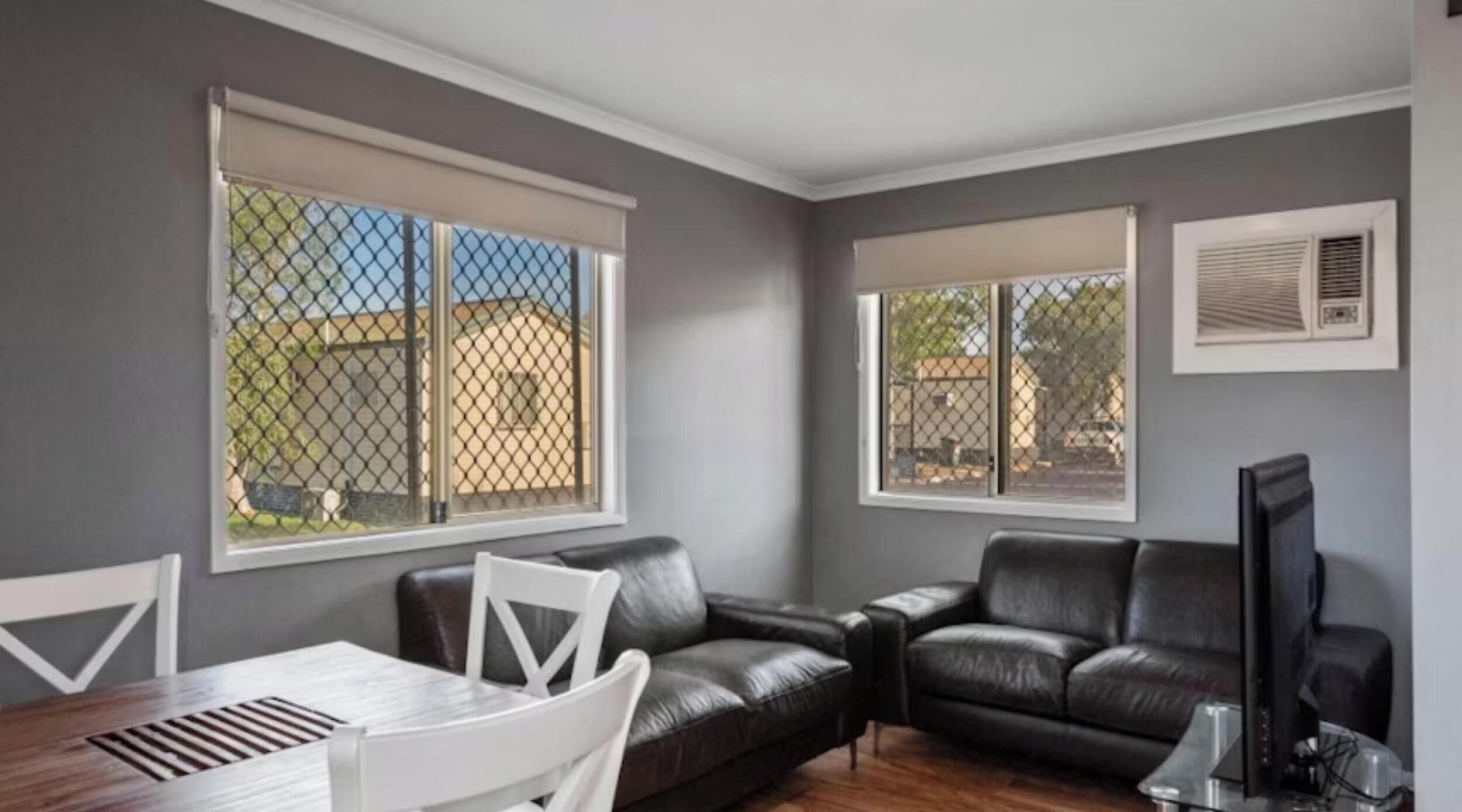 A living room with a grey wall, two black leather sofas, a white dining table, and a TV.