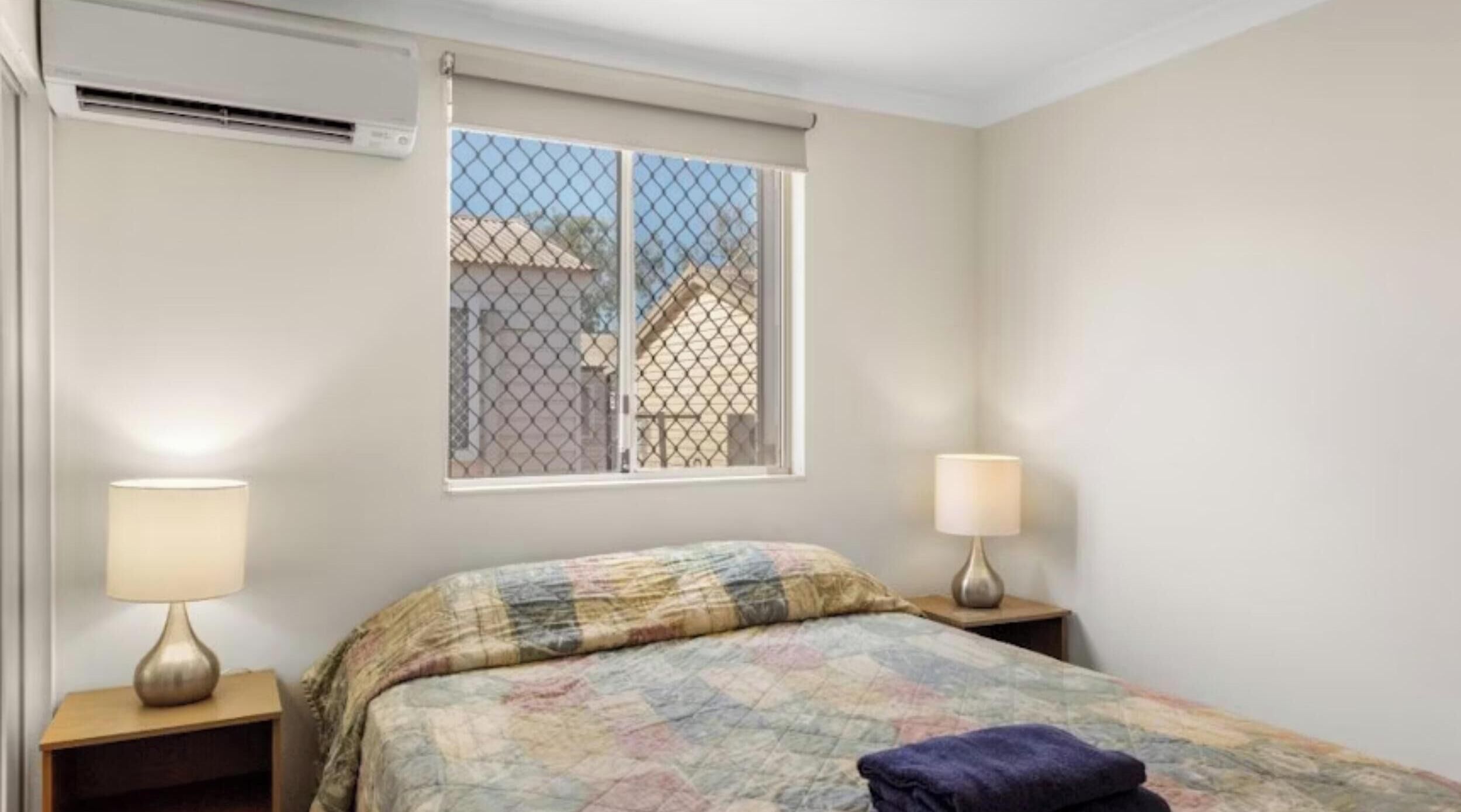 A bedroom with a bed, two bedside lamps, a window with a view, and an air conditioning unit.