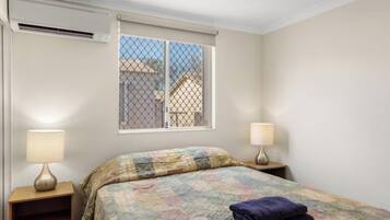 A bedroom with a bed, two bedside lamps, a window with a view, and an air conditioning unit.