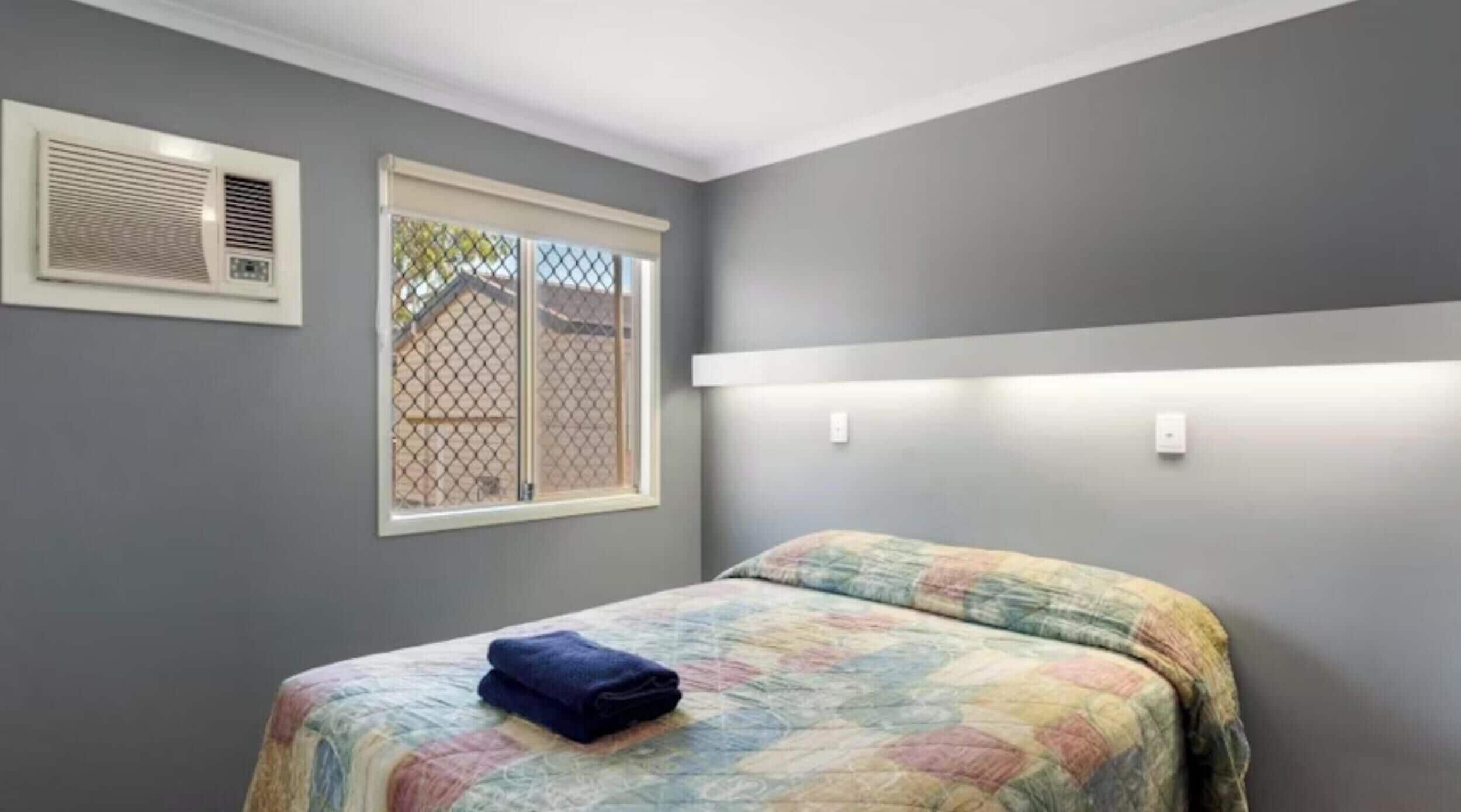 A bedroom with a bed, a window with blinds, and an air conditioning unit.