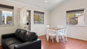Una habitación con un sofá de cuero negro, una mesa de comedor de madera y sillas blancas, y ventanas con persianas.