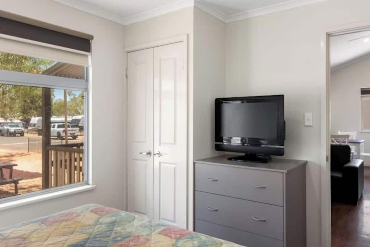 A bedroom with a bed, a television on a dresser, a door, and a window with a view of parked cars outside.