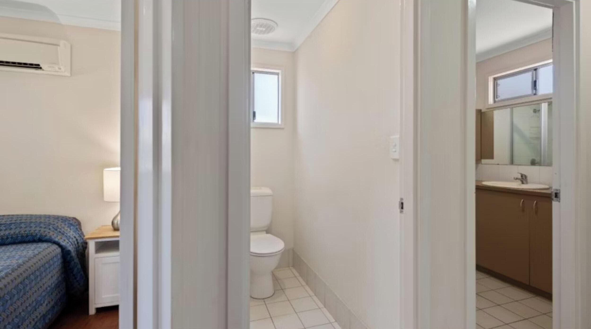A hotel room with a bed, a bedside table, a lamp, a toilet, and a bathroom with a sink and mirror.