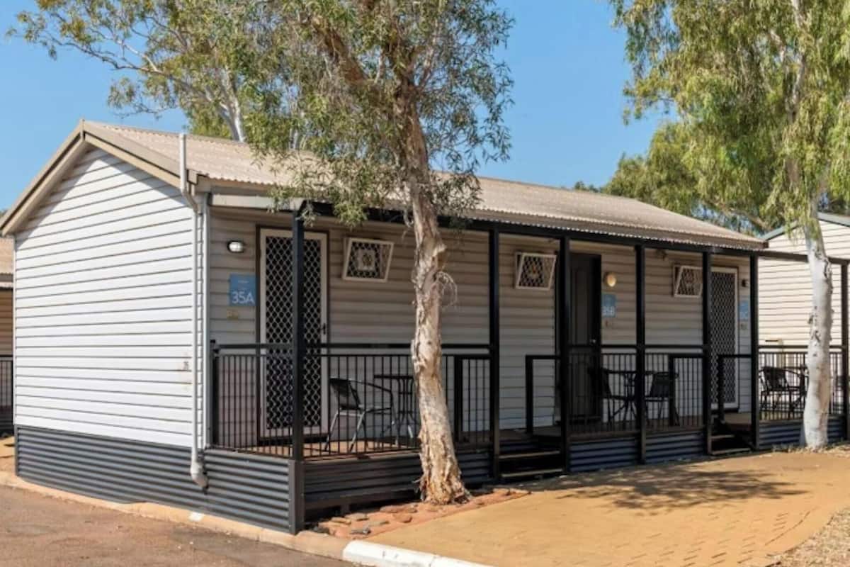 A single-story mobile home with a covered porch, a small outdoor seating area, and a tree in front.