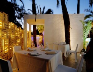 An outdoor dining area with tables set for two, palm trees, and a building with illuminated windows in the background. - Hotel Safari (Gandia)