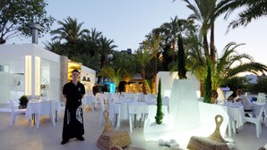 An outdoor dining area with white tables and chairs, a waitstaff member, and a modern building with a glass facade. - Hotel Safari (Gandia)