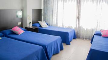 Habitación de hotel con tres camas individuales, cada una con cubrecama azul y almohada rosa. Hay una mesita con una lámpara y una planta en maceta. La habitación tiene un ventanal con cortinas translúcidas y vistas a árboles.