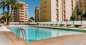 A swimming pool with lounge chairs, surrounded by palm trees and tall buildings. - Hotel Safari (Gandia)