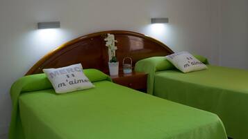 Habitación de hotel con dos camas individuales, cada una con cubrecama verde y almohada con texto. Cuenta con un cabecero de madera, una mesita con un jarrón de flores e iluminación en la pared.