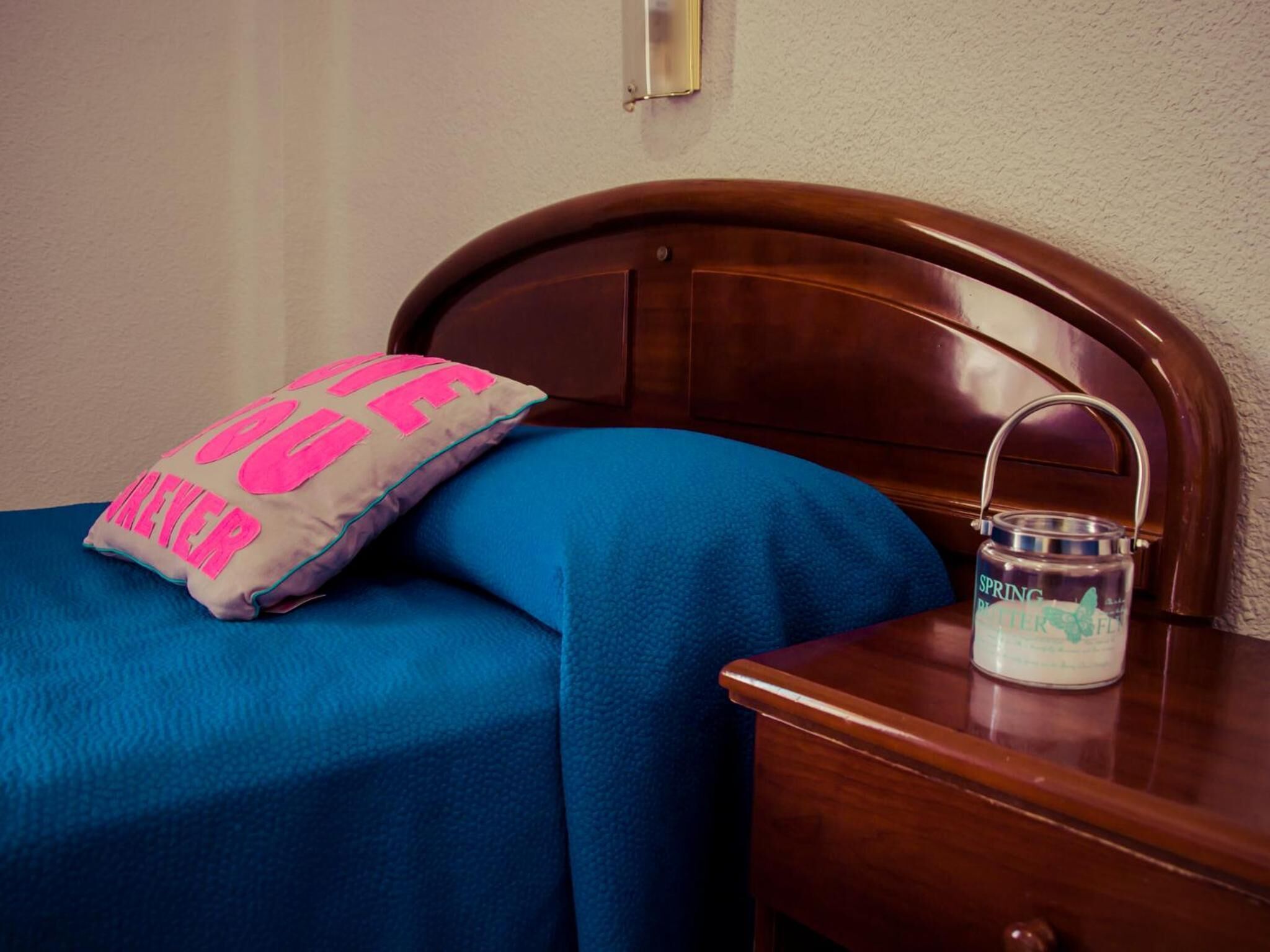 Una cama con colcha azul y almohada con texto rosa, una mesita de noche de madera con un tarro etiquetado 'SPRING BOTTLE FLAVOR'.