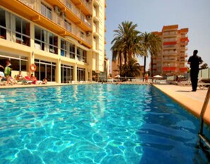 A swimming pool with clear blue water, surrounded by a building with balconies and palm trees. - Hotel Safari (Gandia)