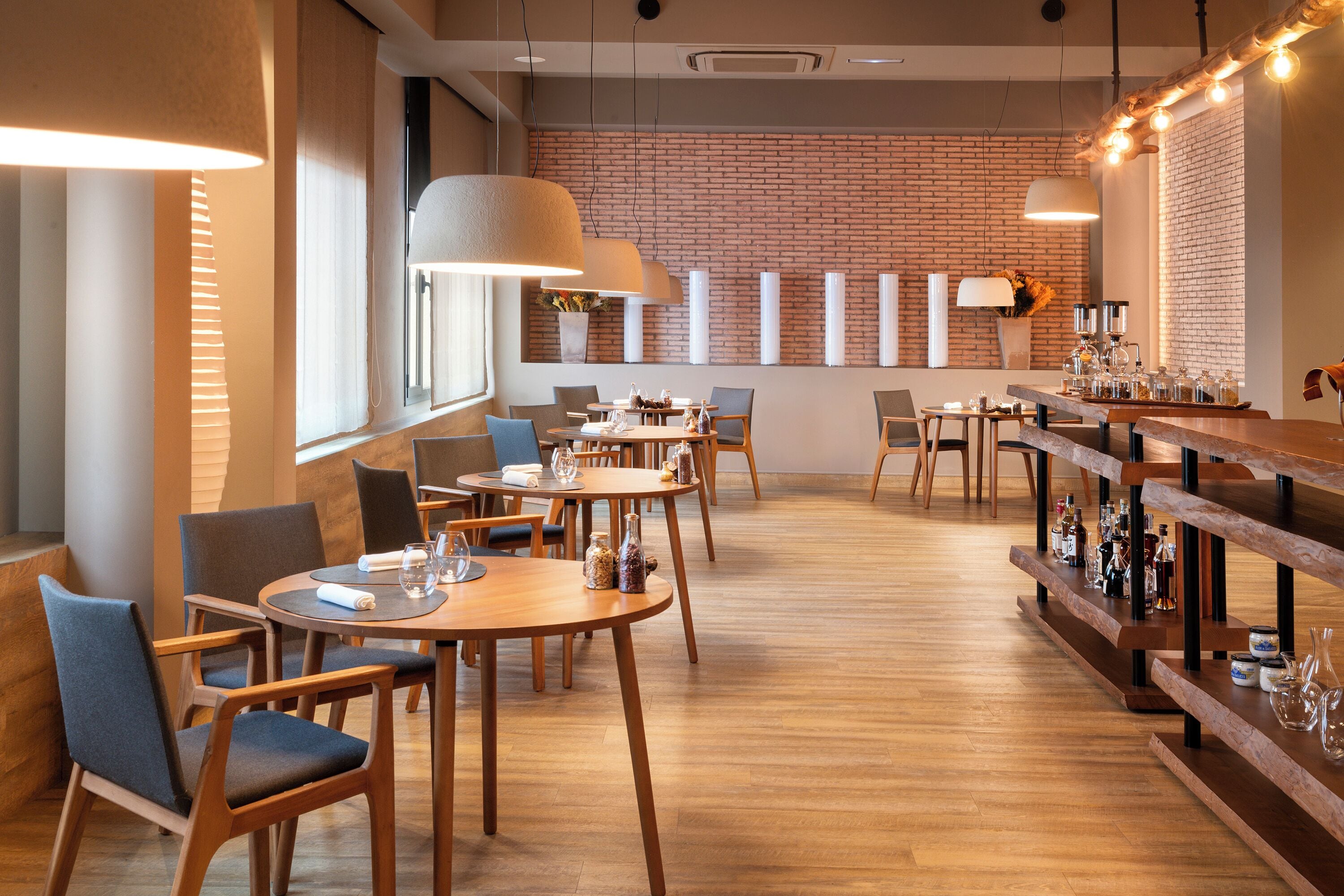 A modern restaurant interior with wooden tables and chairs, a brick wall, and pendant lighting.