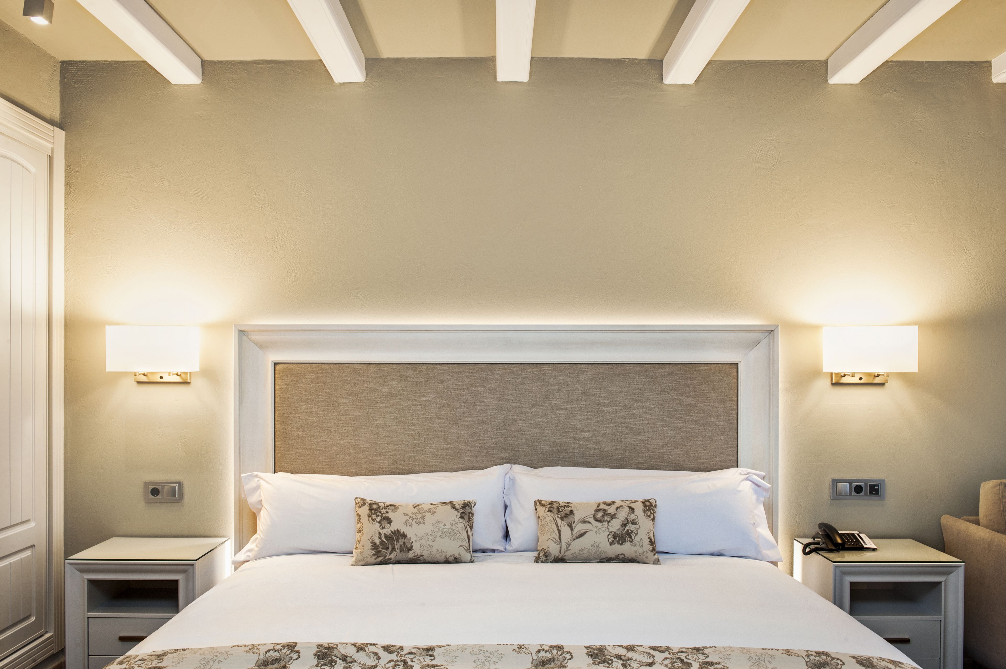 A neatly made bed with floral-patterned pillows, two bedside tables with lamps, and a plain wall.
