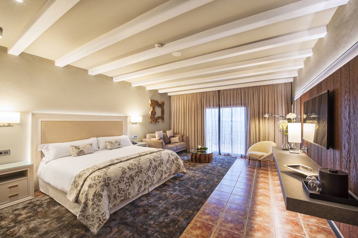 Amplia habitación de hotel con una cama grande, un sofá, una mesita y un televisor fijado en la pared.