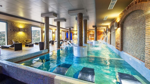 Una piscina interior con agua cristalina, paredes de azulejos y columnas. Se observan sillones de descanso y vistas al exterior.